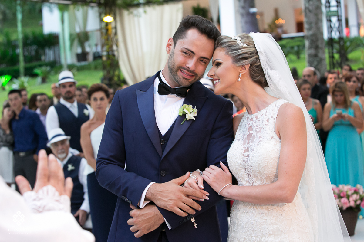 noiva se encosta no ombro do noivo, ambos muito carinhosos, de maos dadas, ao fundo se ve o cenário da nave onde acontecem as cerimônas de casamento na fazenda 7 lagoas.