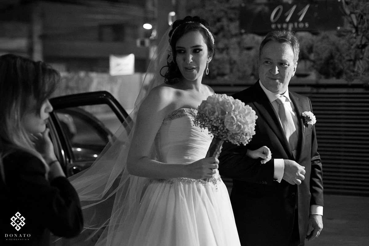Noiva saindo do carro com seu pai, ao fundo da pra ver o logo do buffet 011 eventos.