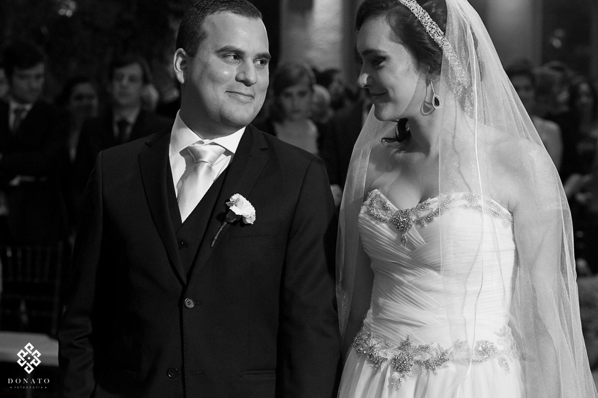 noivos se olham no altar e trocam sorrisos, a foto esta em branco e preto, dando assim um ar mais romántico a cena.