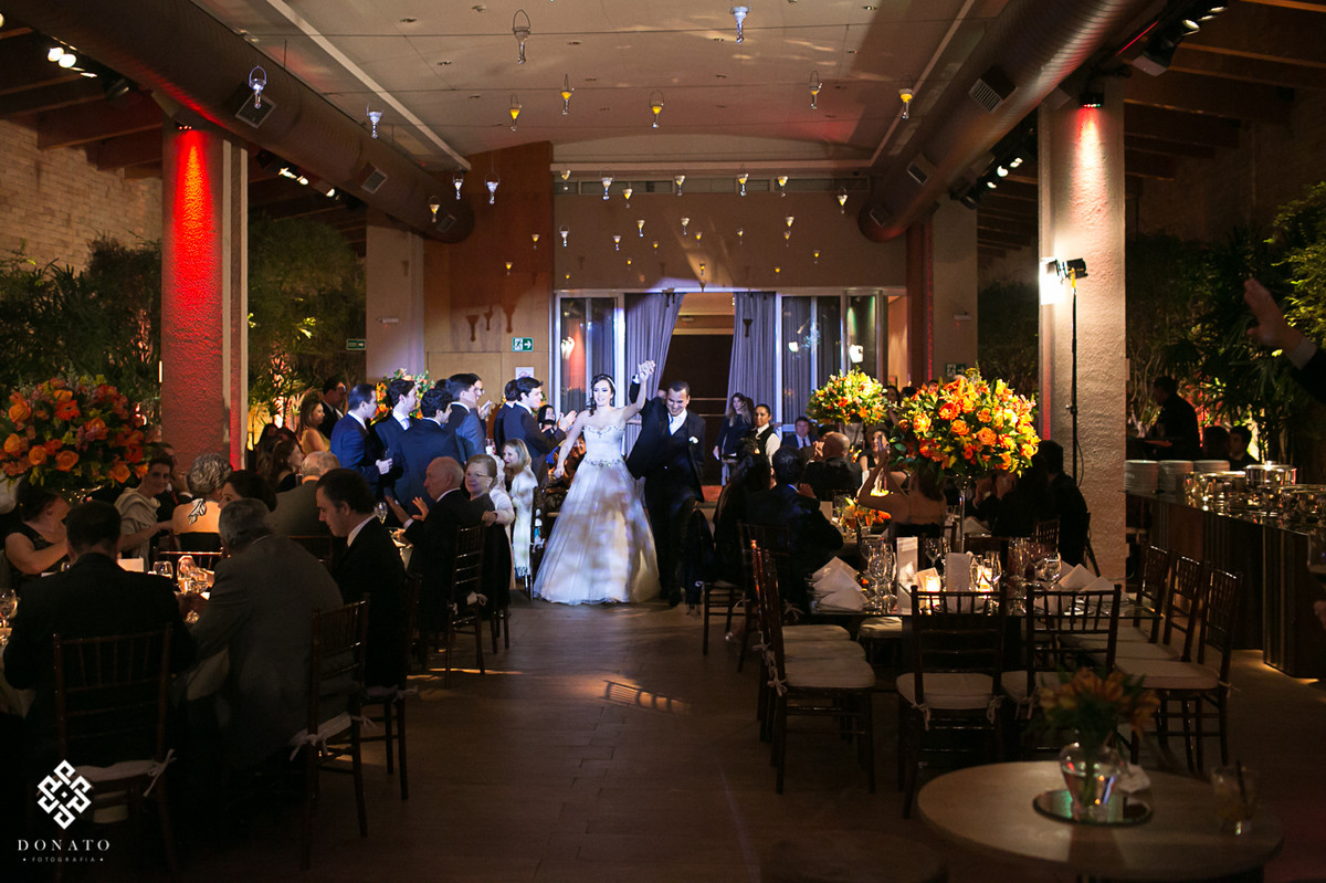 noivos entram na festa, foto muito alegre, a noiva levanta o braço comemorando, e todos os convidados comemoram juntos.