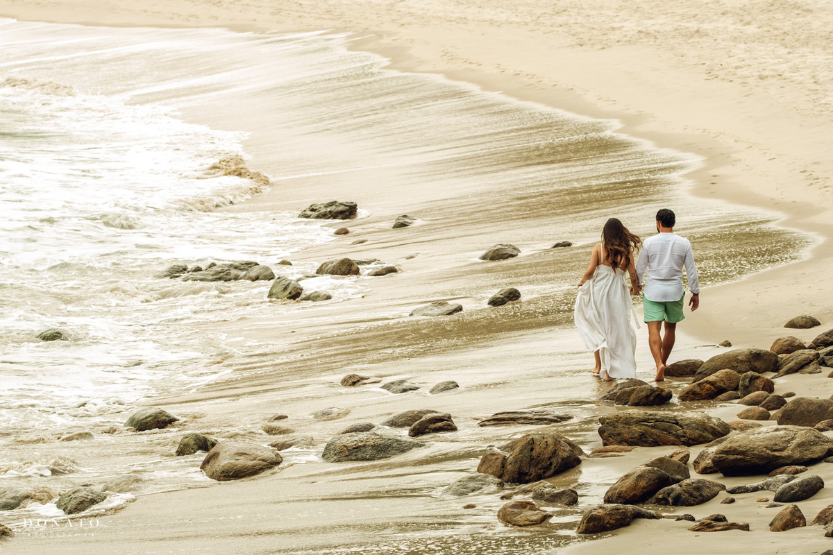 Foto dos noivos caminhando na praia do Guarujá, de mãos dadas com o mar ao fundo.
