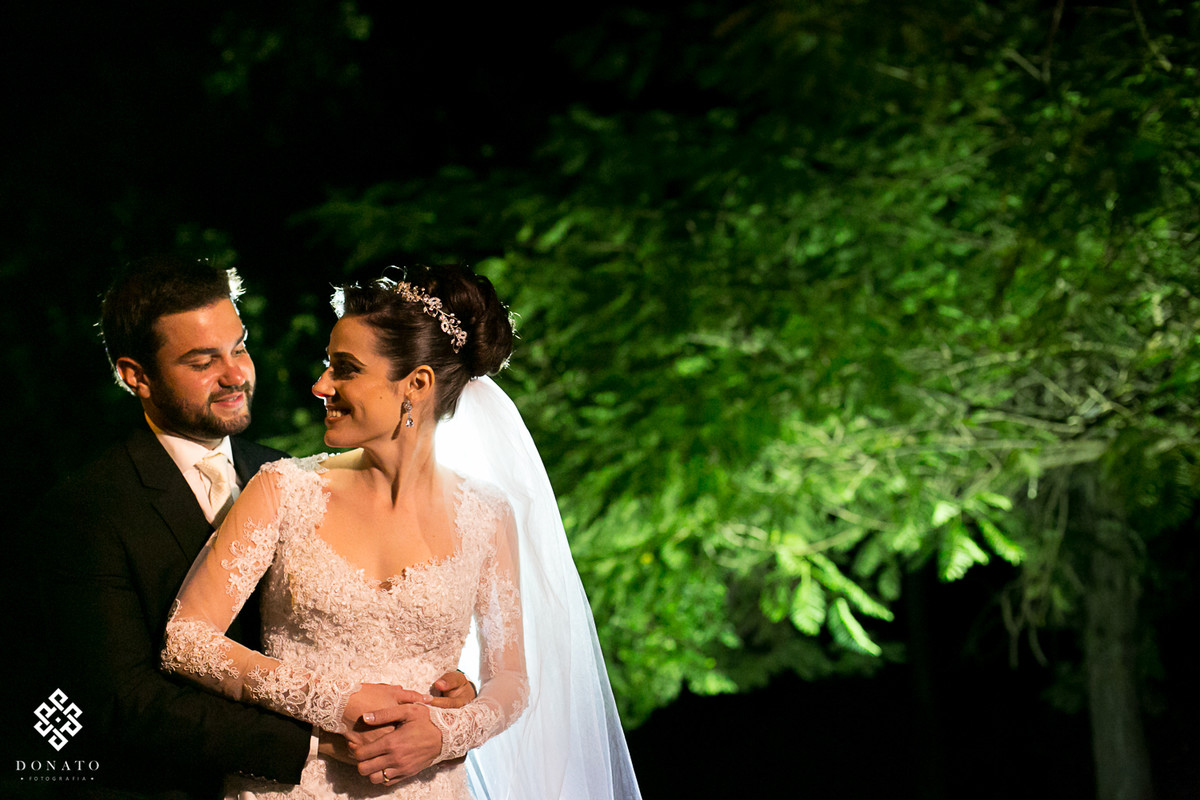 Foto feita de baixo de uma arvore linda, que fica no jardim do buffet Flamboyant, a arvore esta toda verde ao fundo e os noivos com um tom quente que remete a paixão.