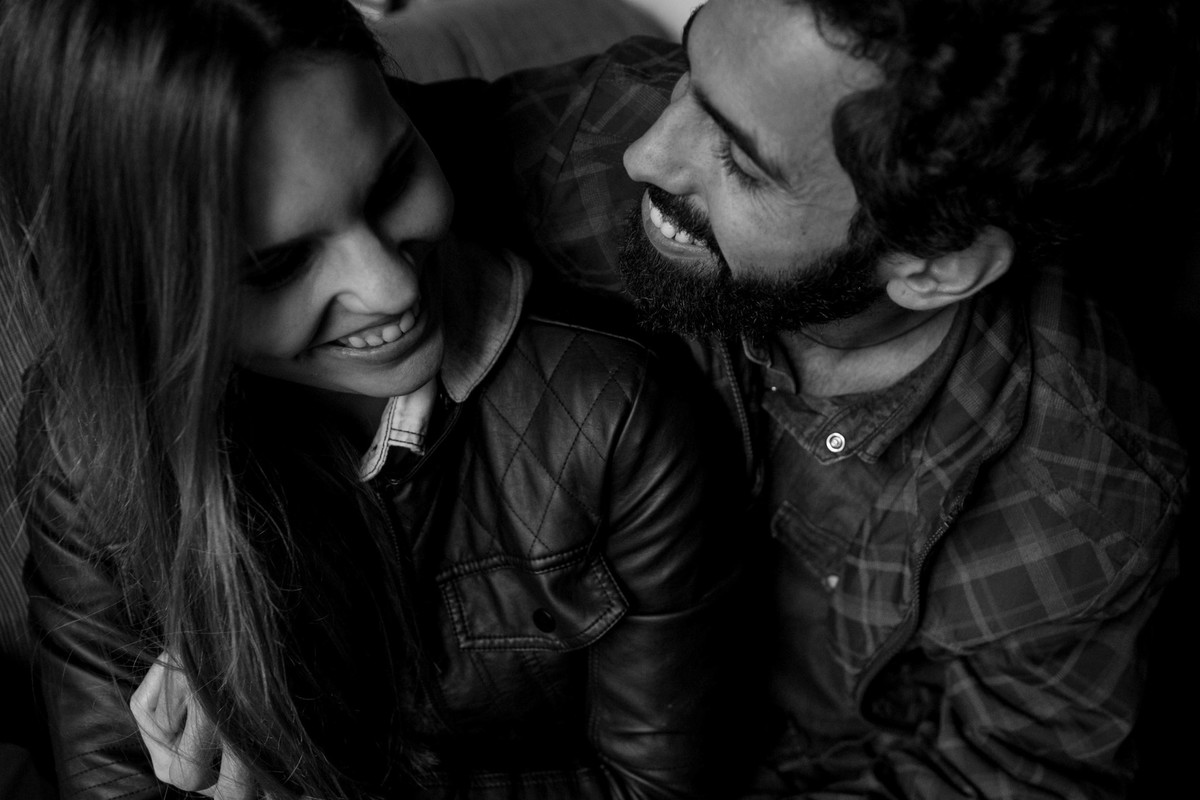 foto bem divertida do casal se olhando e rindo espontaneamente, com uma luz lateral linda vindo de uma porta, para deixar a foto mais linda ela foi feita em branco e preto.