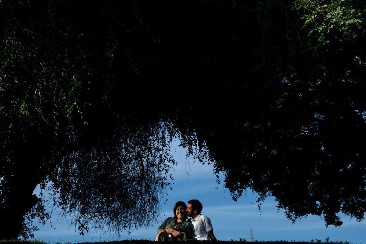 foto do casal sentados entre uma molduras de arvores, ao fundo um lindo céu azul.