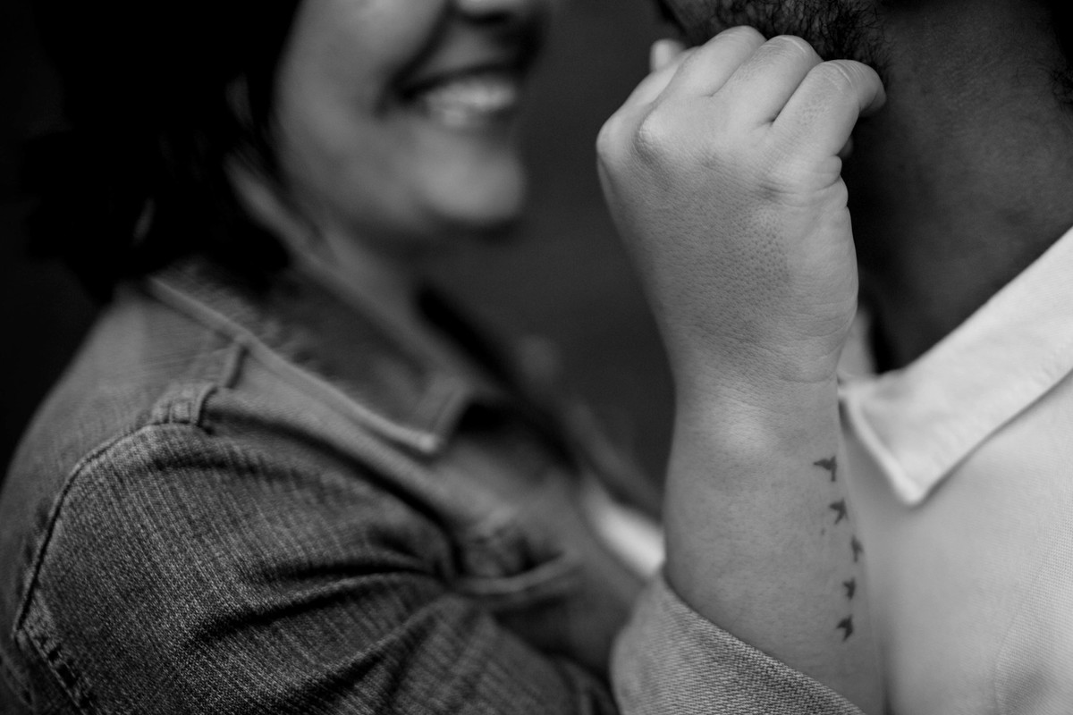 foto do detalhe da tatuagem do braço da noiva, com o lindo sorriso do casal ao fundo em desfoque.