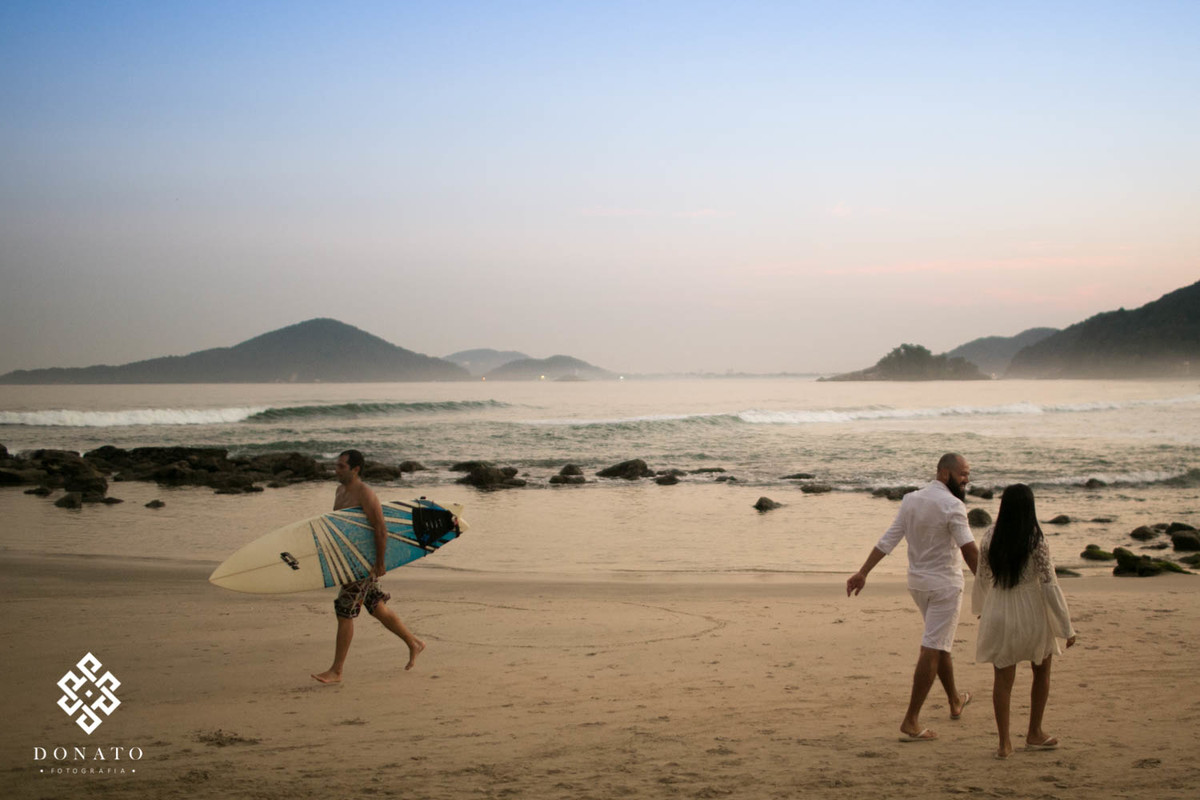 casal caminha para direita, enquanto surfista caminha para esquerda, ao fundo o mar e as montanhas, deixando assim a imagem mais bonita de se ver.