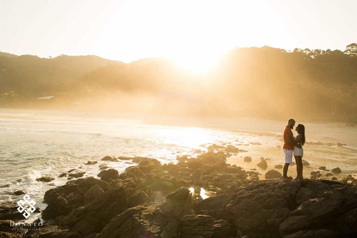 imagem da praia ao fundo, com uma luz dourado do sol, o casal namorando em pé nas pedras a direita da imagem, linda foto.