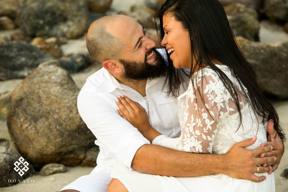 Casal se diverte sentados sobre as pedras na areia da praia, ele esta de camisa branca e ela com vestido branco de rendinha.