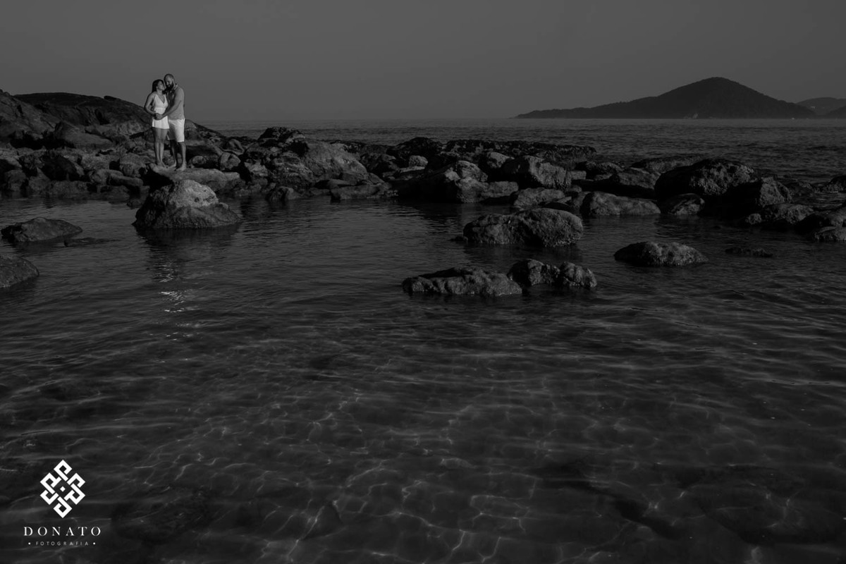 imagem da praia e as pedras, com o casal pequeno ao fundo, a agua cristalina e transparente reflete o fundo, tudo isso foi feito em branco e preto.