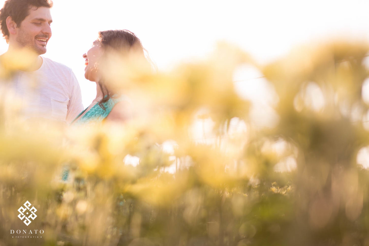 Casal se diverte entre margaridas amarelas, as flores estão borradas.