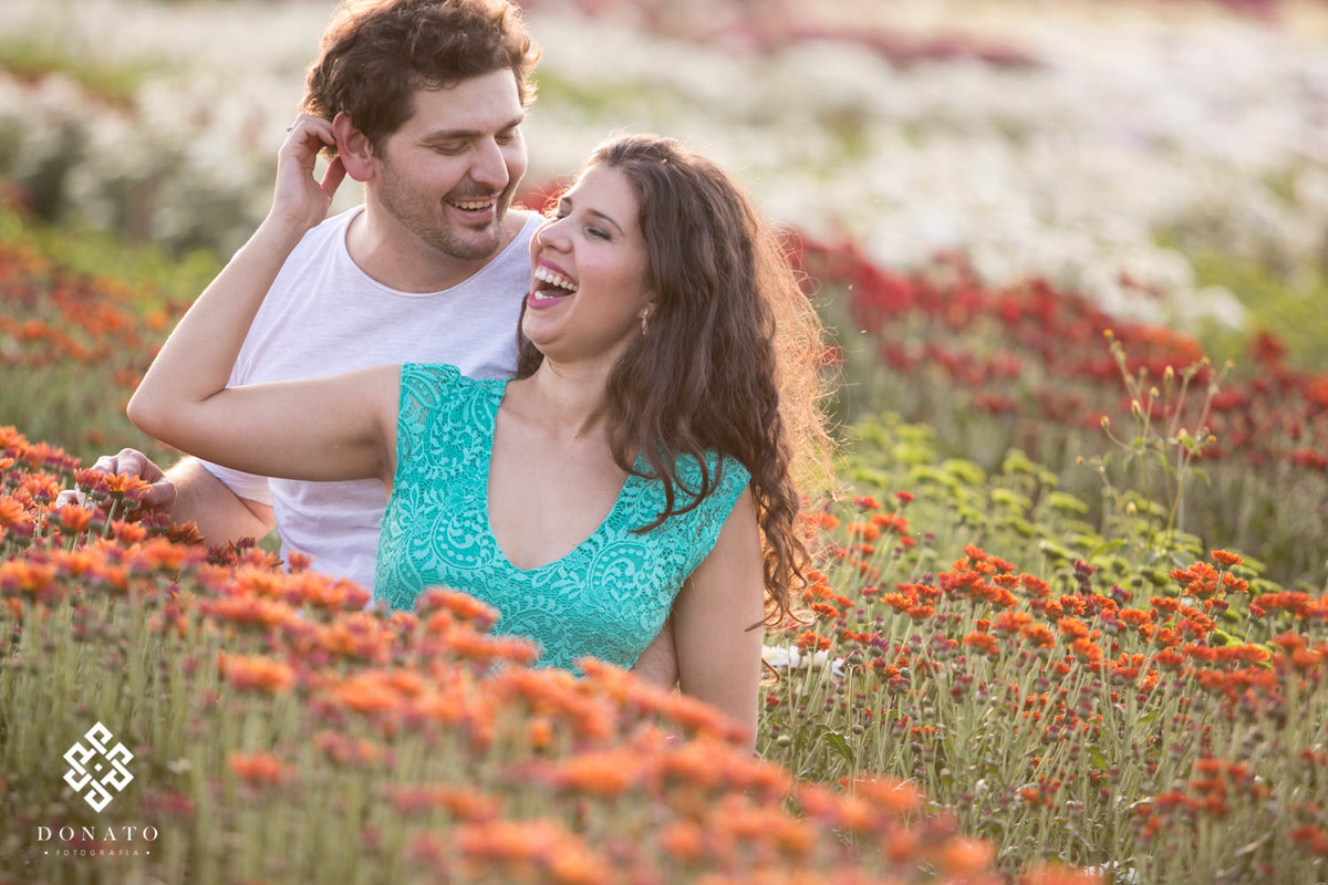 Um sorriso muito bonito do casal, a foto foi feita em diagonal, formando linhas laterais de flores laranja, verdes, vermelhas e brancas ao fundo.