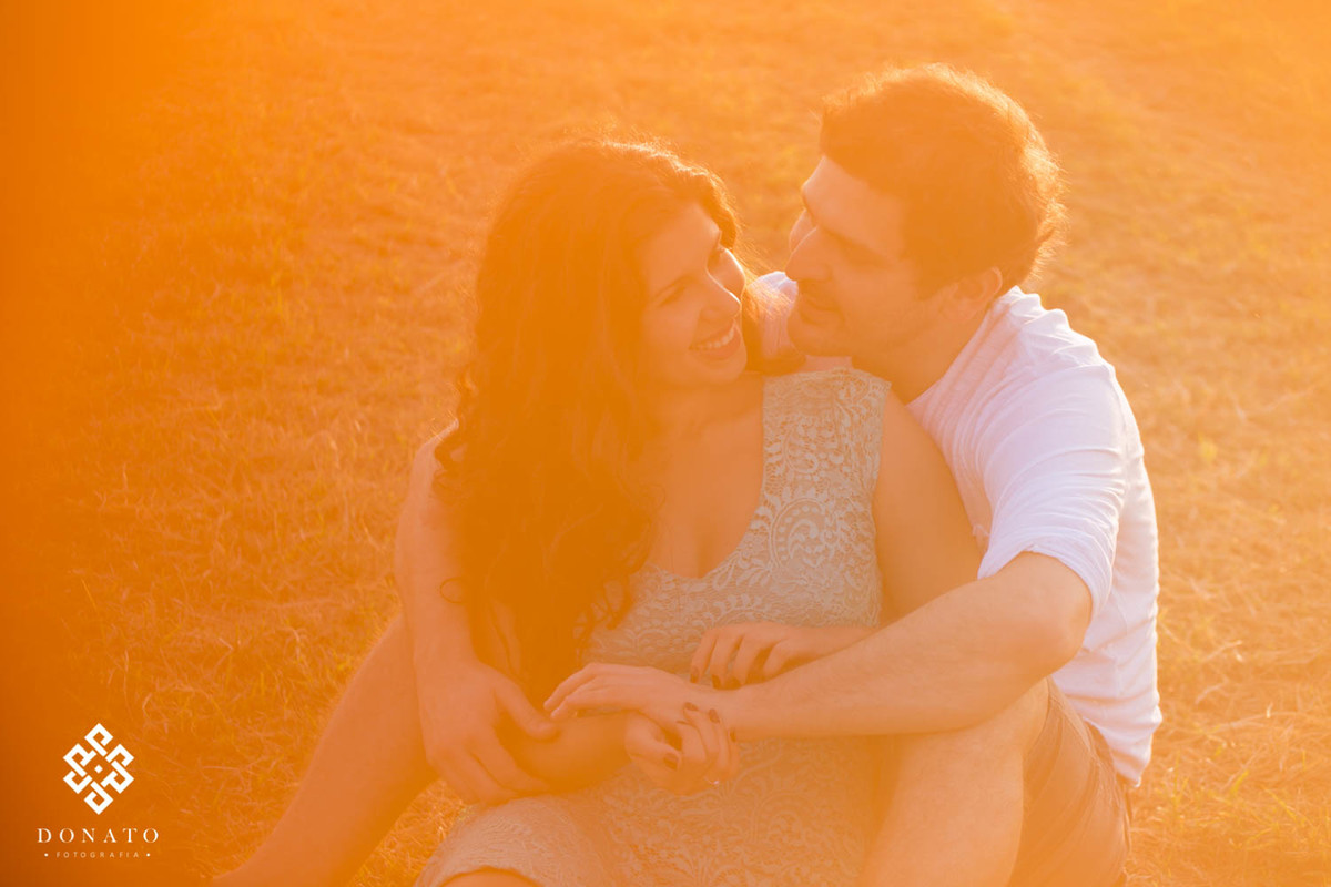 Noivos namoram sentados no feno ainda baixo, uma luz alaranjada entra em toda a foto, deixando um ar ainda mais romântico a foto.