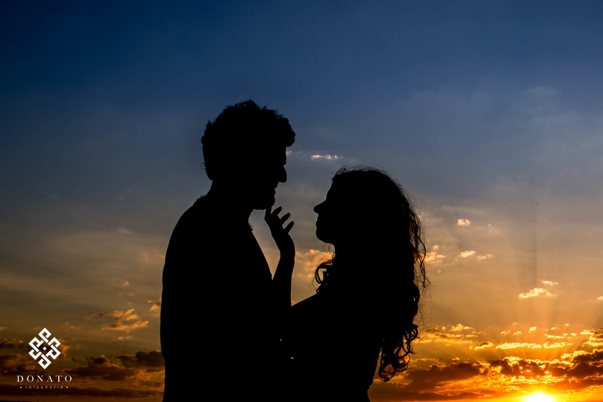 Uma foto com um lindo por do sol, o casal em silhueta, a noiva faz um carinho delicado na barba do noivo.