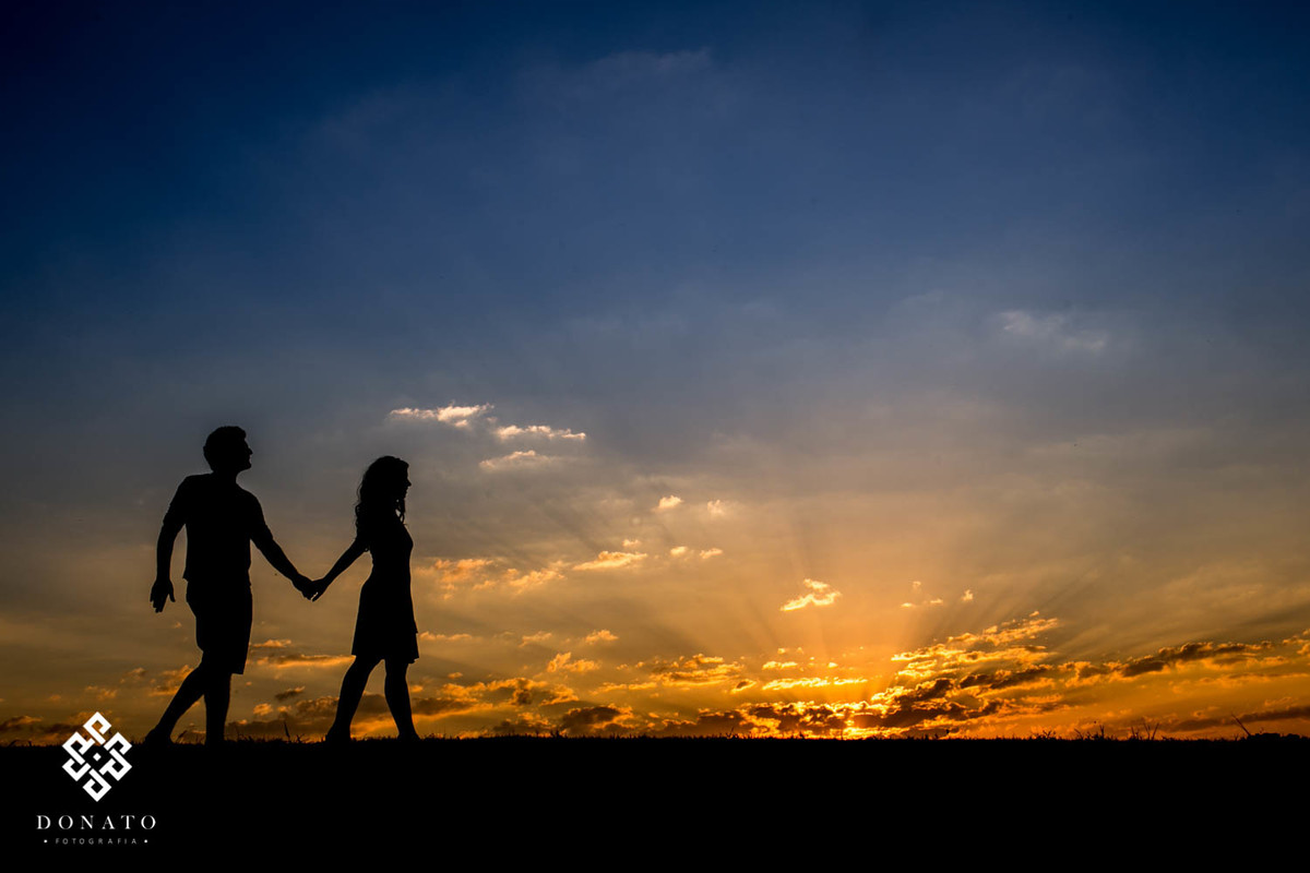 noivos caminham de maos dadas em frente a um lindo por do sol, a imagem tem um ceu azul maravilhoso e a pate de baixo toda dourada do sol.