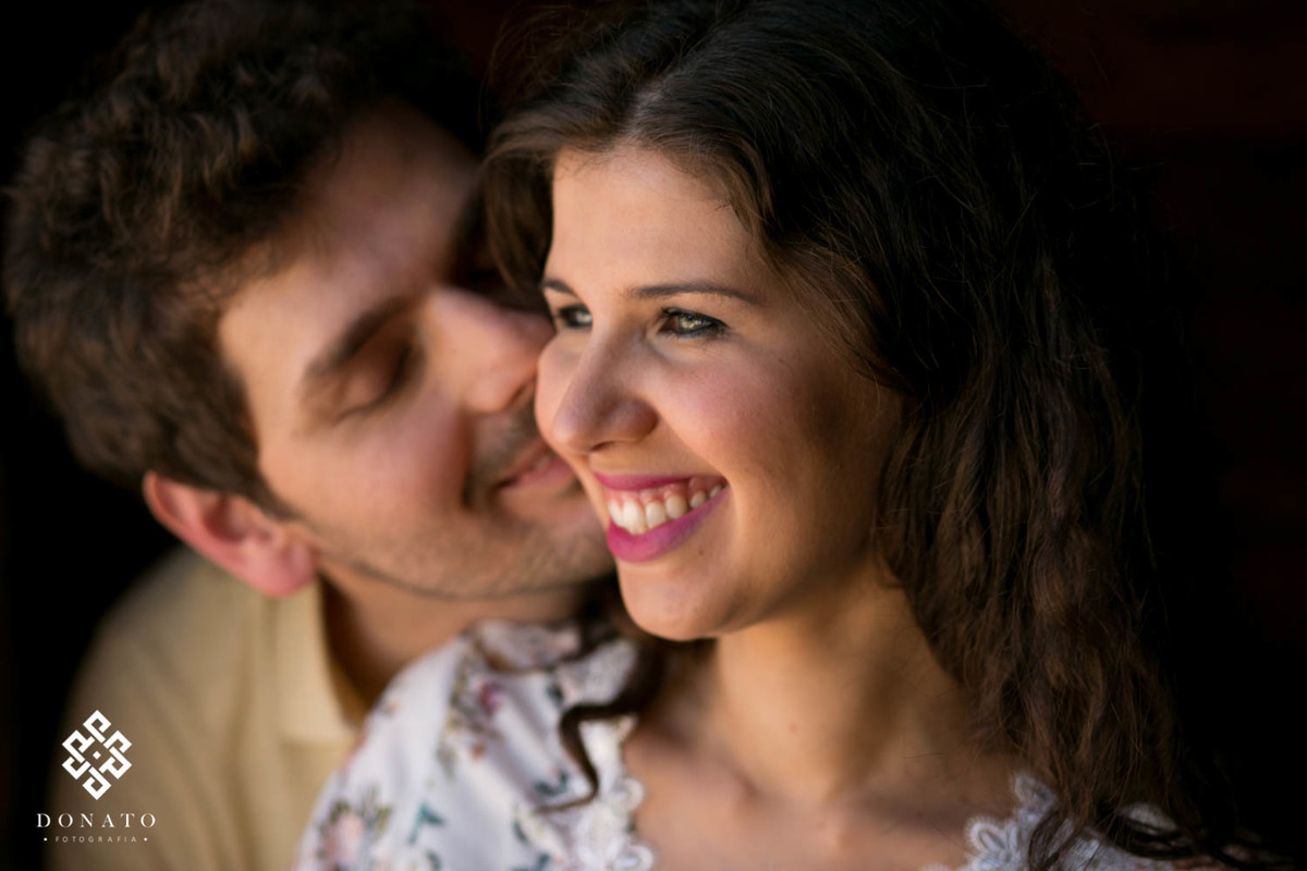 A noiva sorri com seu lindos olhos verdes, enquanto o noivo da um cheiro no pescoço, a luz é suave e a imagem encantadora.