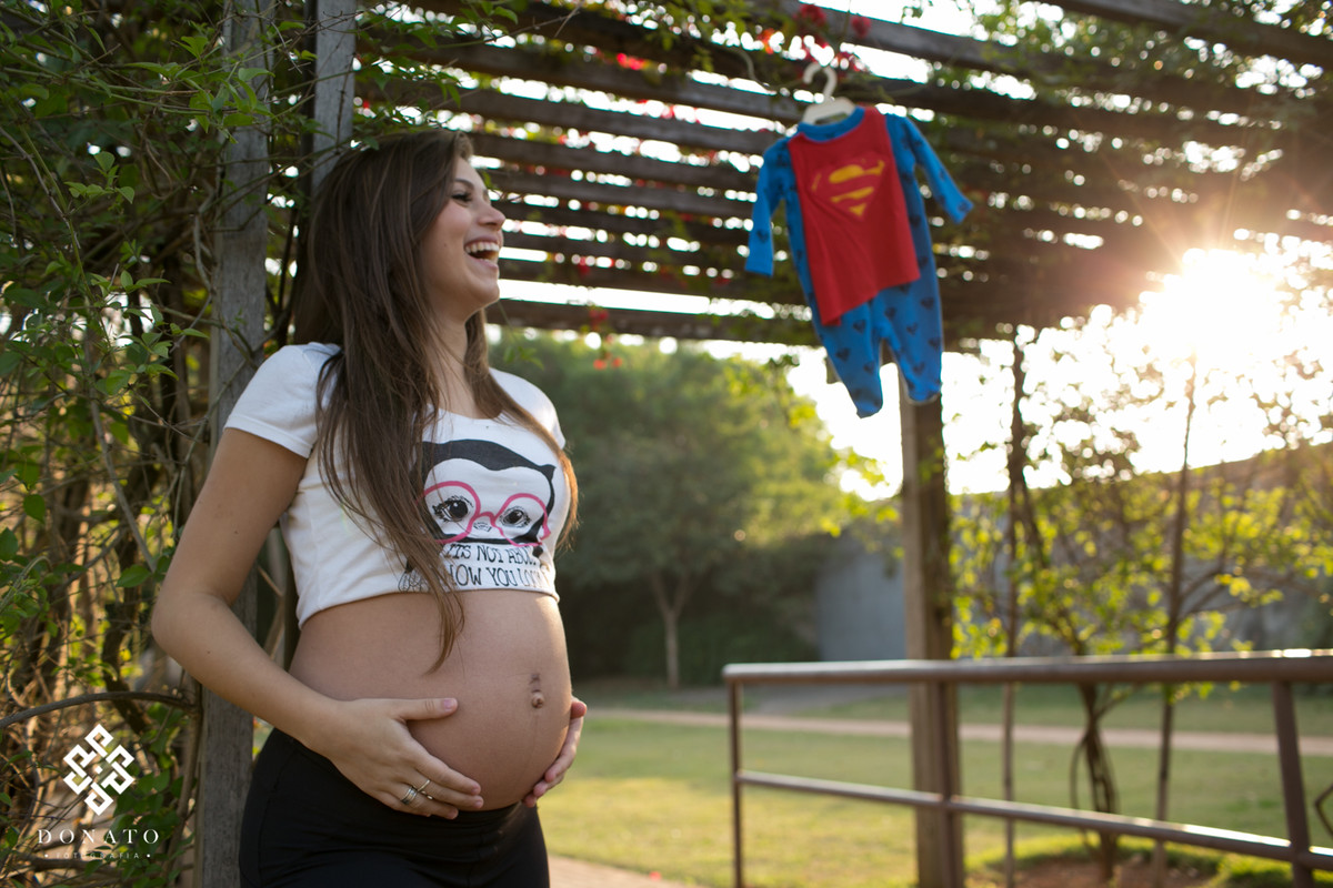 a gestante leticia mostrando a barriga,  ela esta dando um sorriso lindo, ao fundo a roupinha de super homem do nenem pendurada