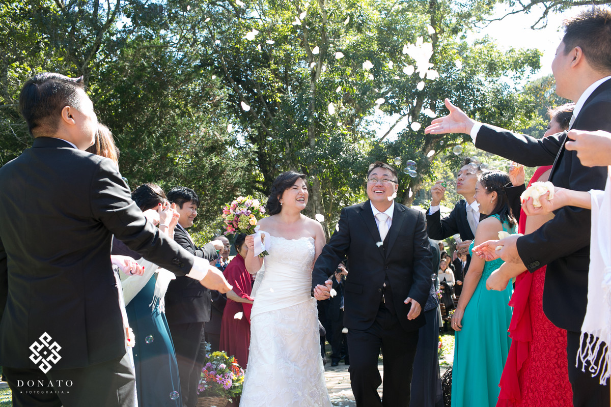 saida dos noivos da cerimonia, os padrinhos jogam pétalas de flores, os noivos sorriem, o casamento foi durante o dia, o que deixou a imagem ainda mais bonita.
