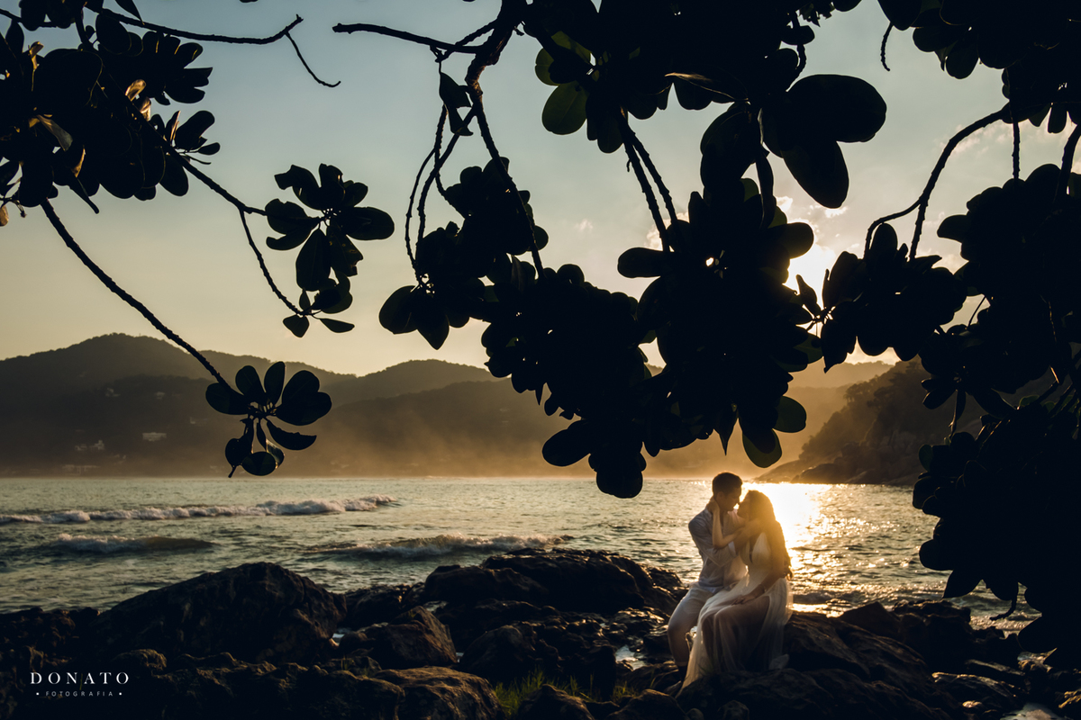 casal em pre-wedding sentados nas pedras da praia das conchas no Guarujá com um lindo por do sol.