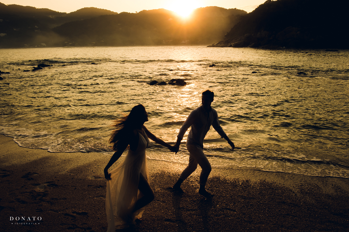 noivos correm na praia com por do sol ao fundo.