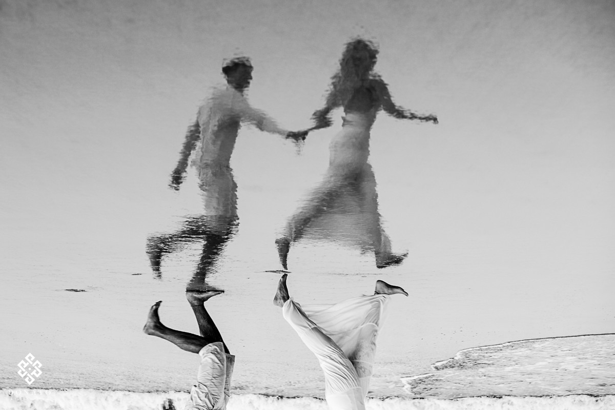 noivos correm de mãos dadas na agua do mar, a imagem é refletida na areia da praia, a foto é incrível.