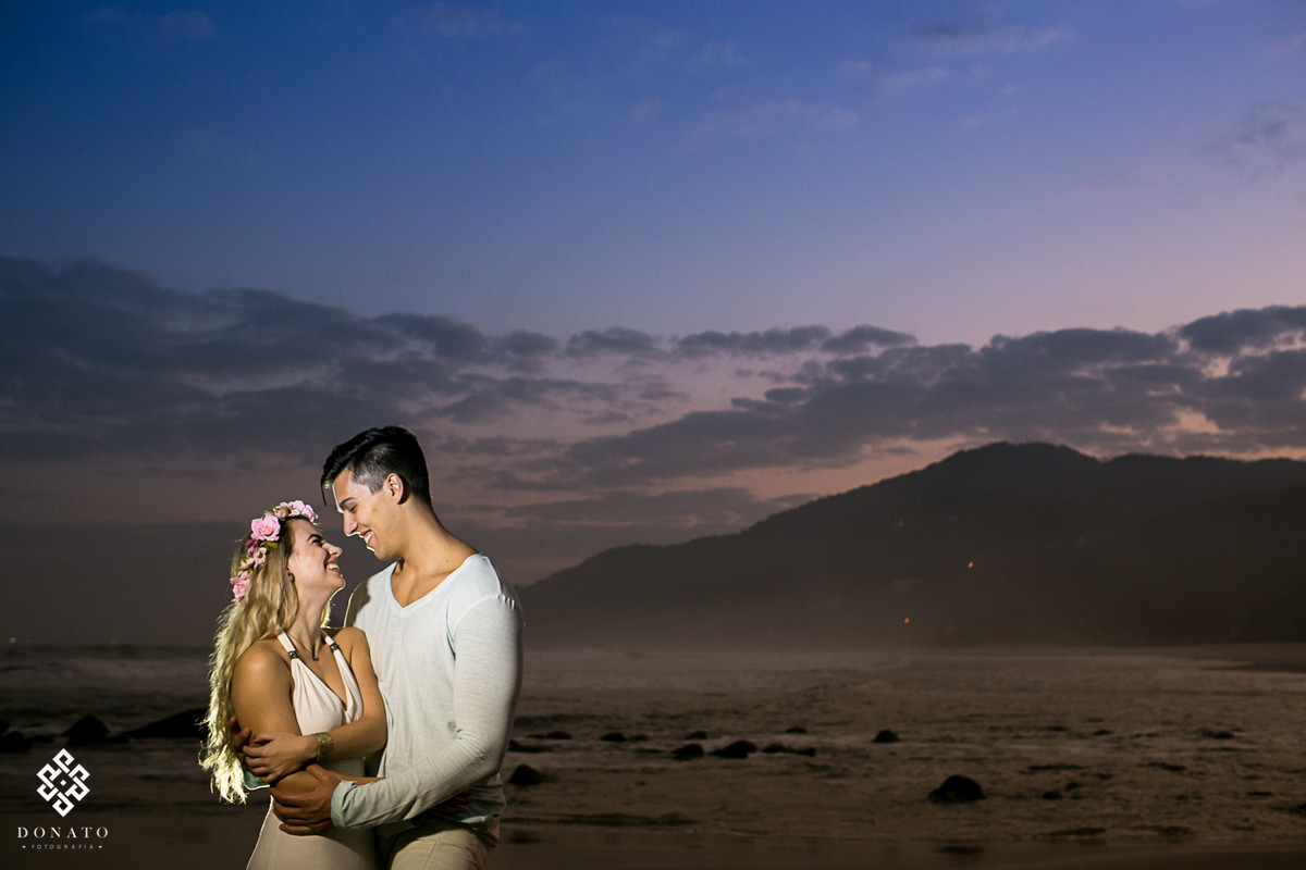 foto do pre wedding, com noivos abraçados na praia e o por do sol ao fundo.