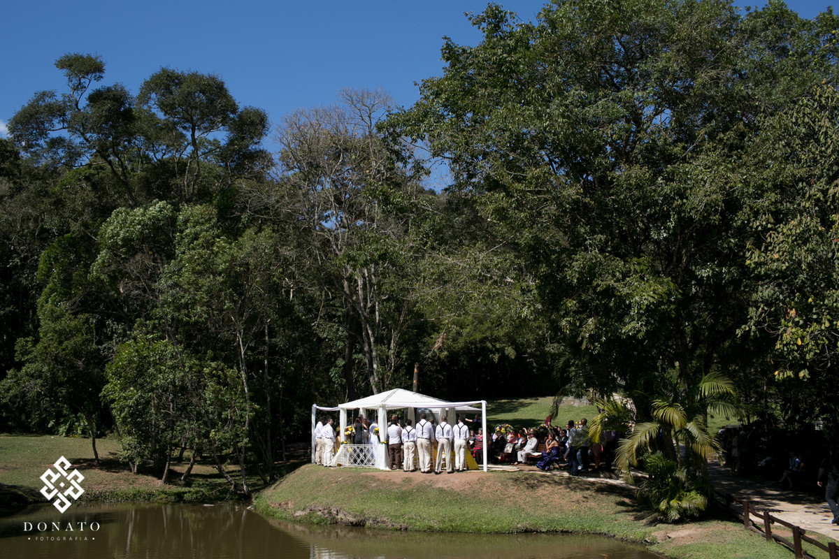 Visão geral da cerimonia do espaço natureza, com todos os padrinhos perfilados, na paisagem tem um gazebo todo branco entre grandes arvores e um lindo lago.