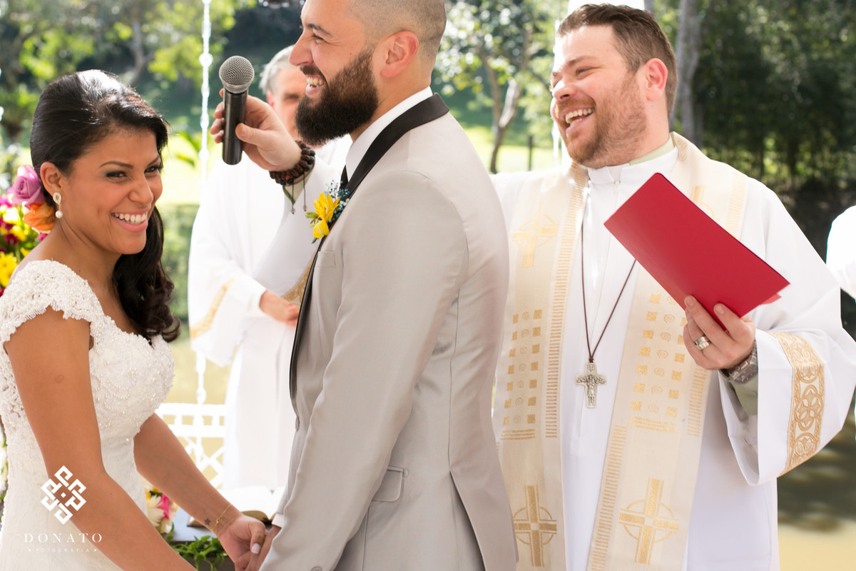 Noivos dao risada, eles estão se olhando ao fundo o reverendo lucas também ri da situação.