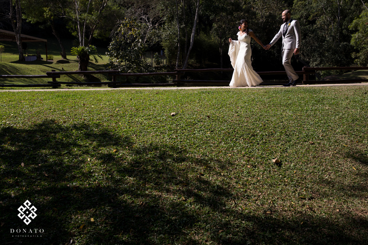 Noivos caminham em frente ao lago, com destino a festa, o lago do espaço natureza ao fundo da a imagem um charme.
