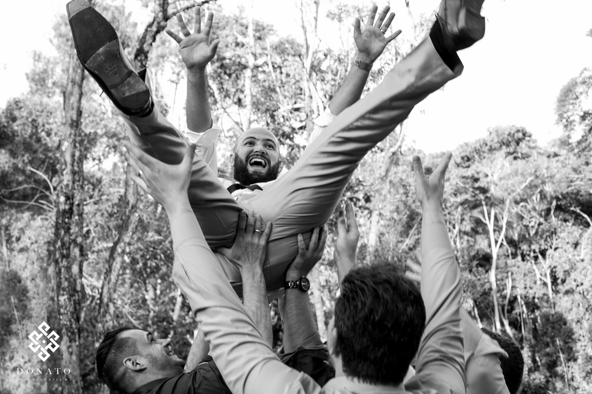 Agora os convidados jogam o noivo pra cima, ele esta de pernas abertas e braços abertos, com um lindo sorriso no rosto, ao fundo arvores.