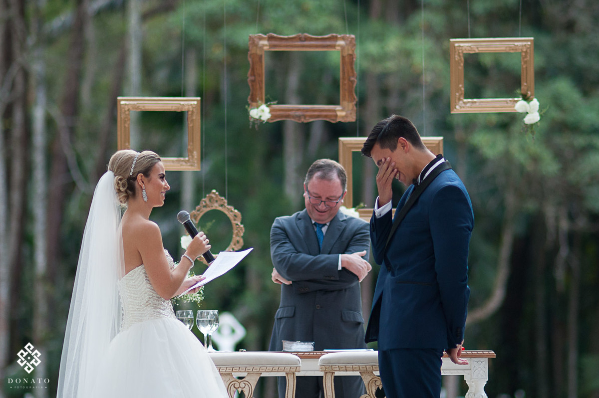 noivo jora emocionado nos votos, a noiva esta falando no microfone e o pastor rindo atrás.