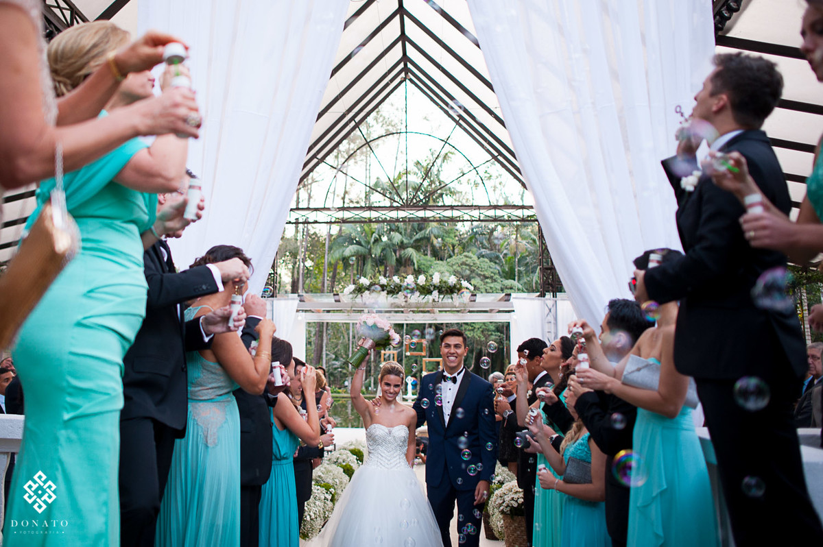 Noiva sai e comemora com o buquet levantado, o padrinhos soltam bolinhas de sabão.