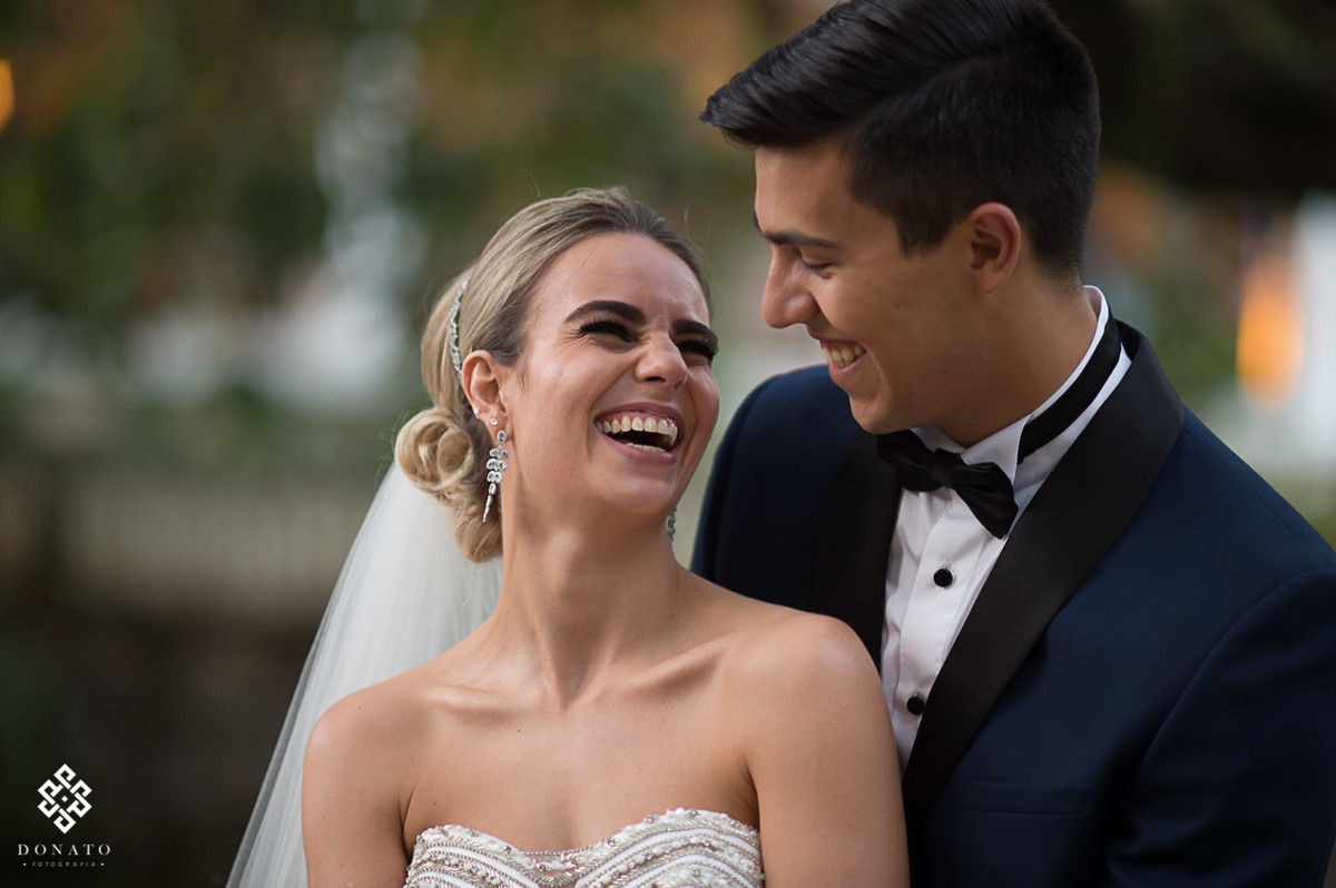 noiva se diverte e da um largo sorriso, enquanto é abraçada pelo noivo.