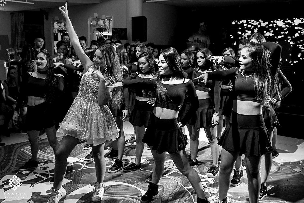 amigas dançam junto com a debutante, ela lidera a dança a frente, ela esta de branco e as amigas de preto.