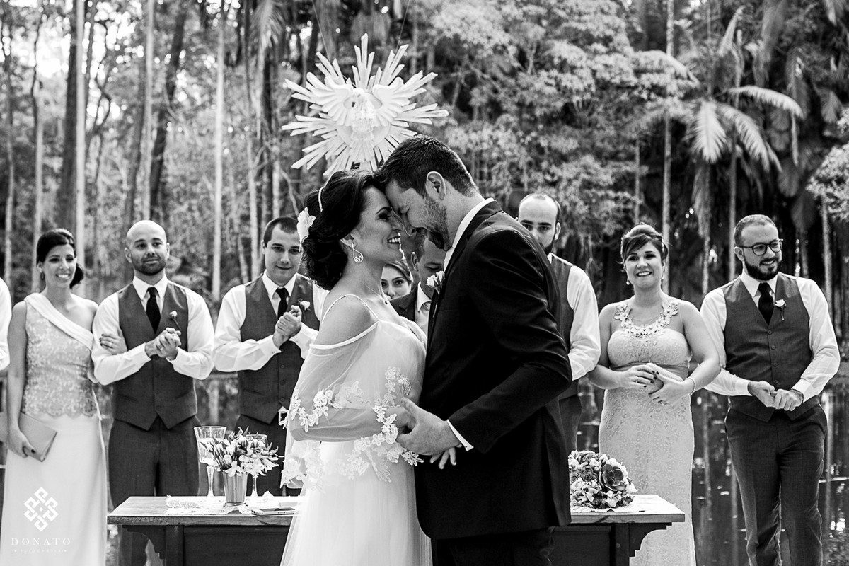 noivos encostam as cabeças e se curtem, ao fundo os padrinhos todos emocionados.