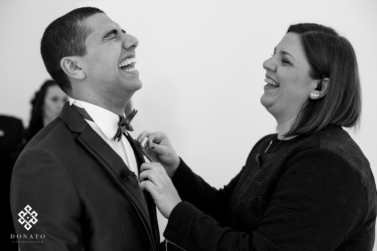 assessora arruma a gravata do noivo, ambos dão uma gargalhada muito divertida.