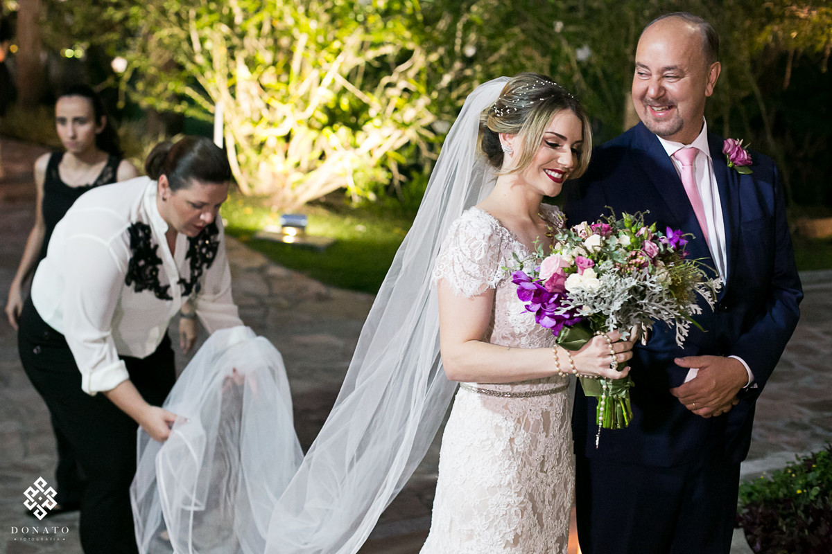 Noiva e pai, se preparam para entrada enquanto a assessora da na barra do meu vestido arruma a cauda do vestido ca noiva.