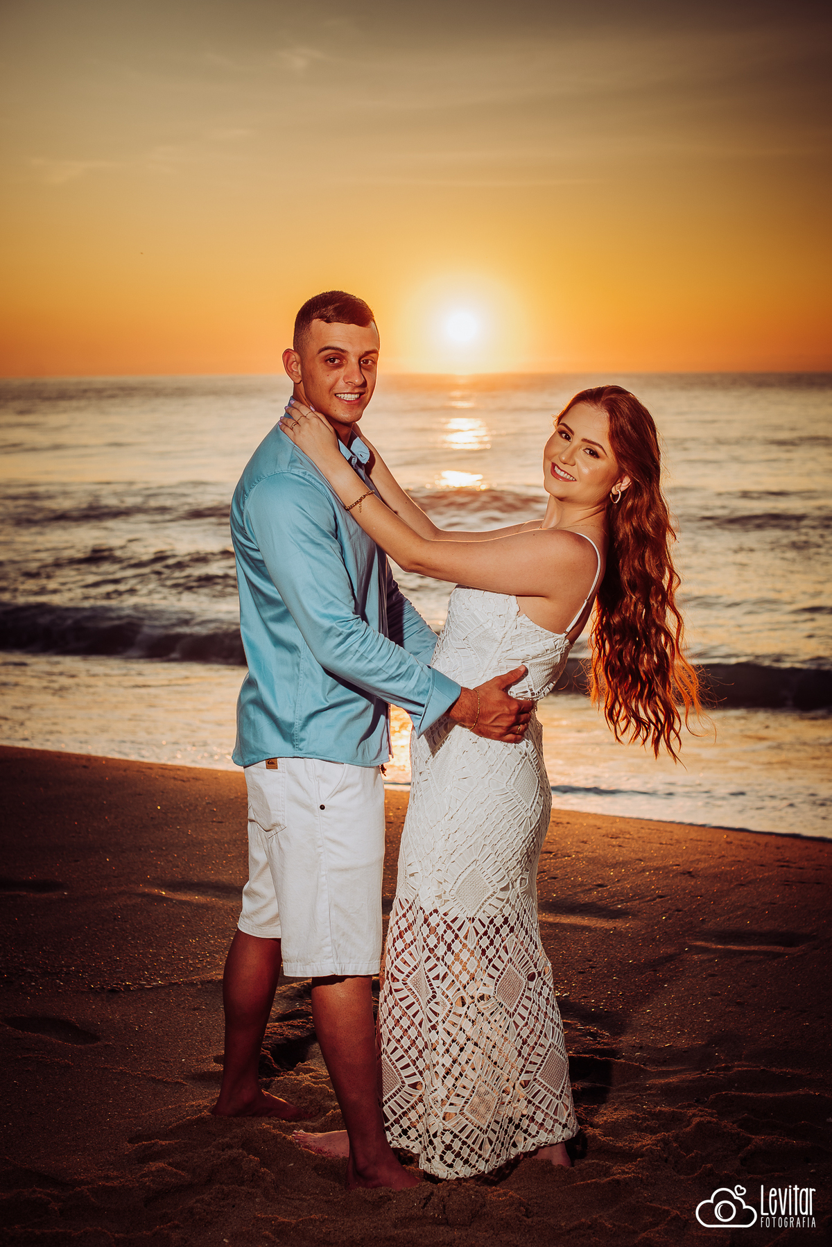 noivos olhando para foto com o nascer do sol refletindo no mar