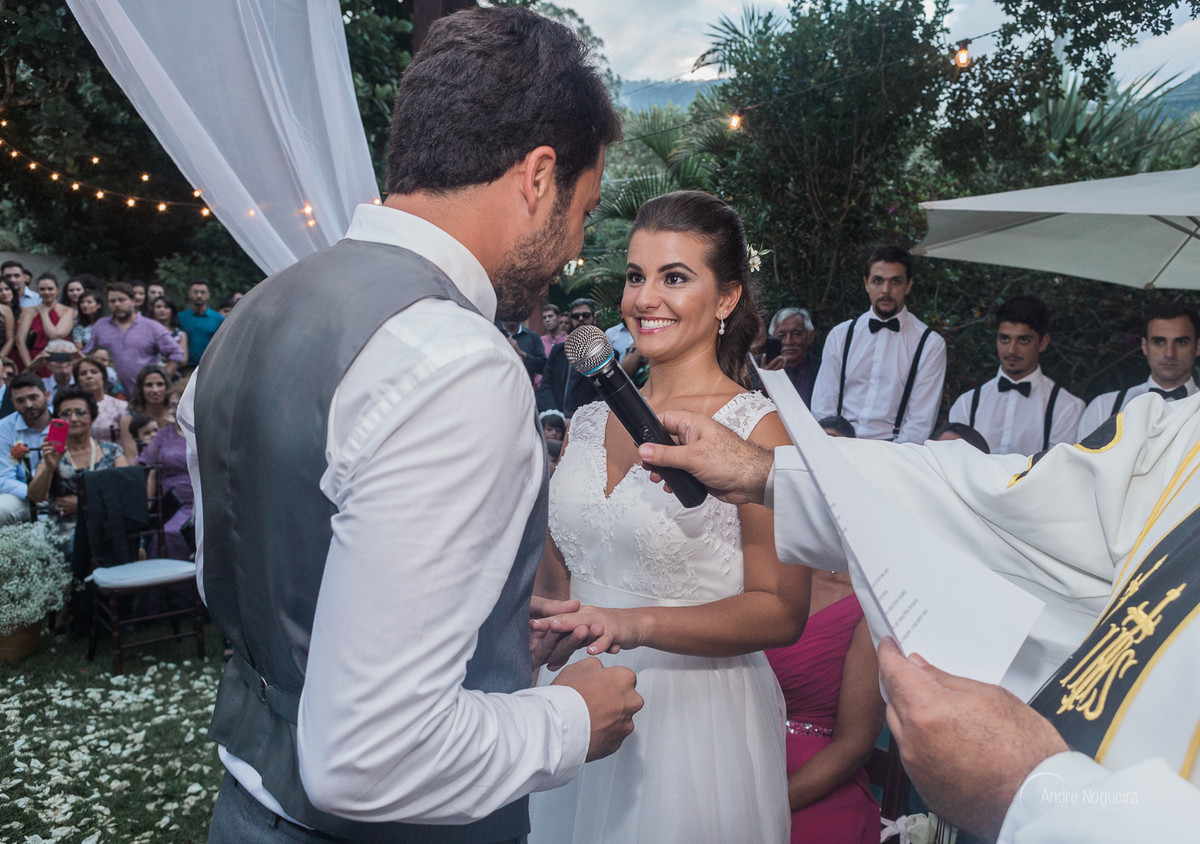 no ritual das alianças oo noivo coloca a aliança no dedo da noiva que observa sorridente