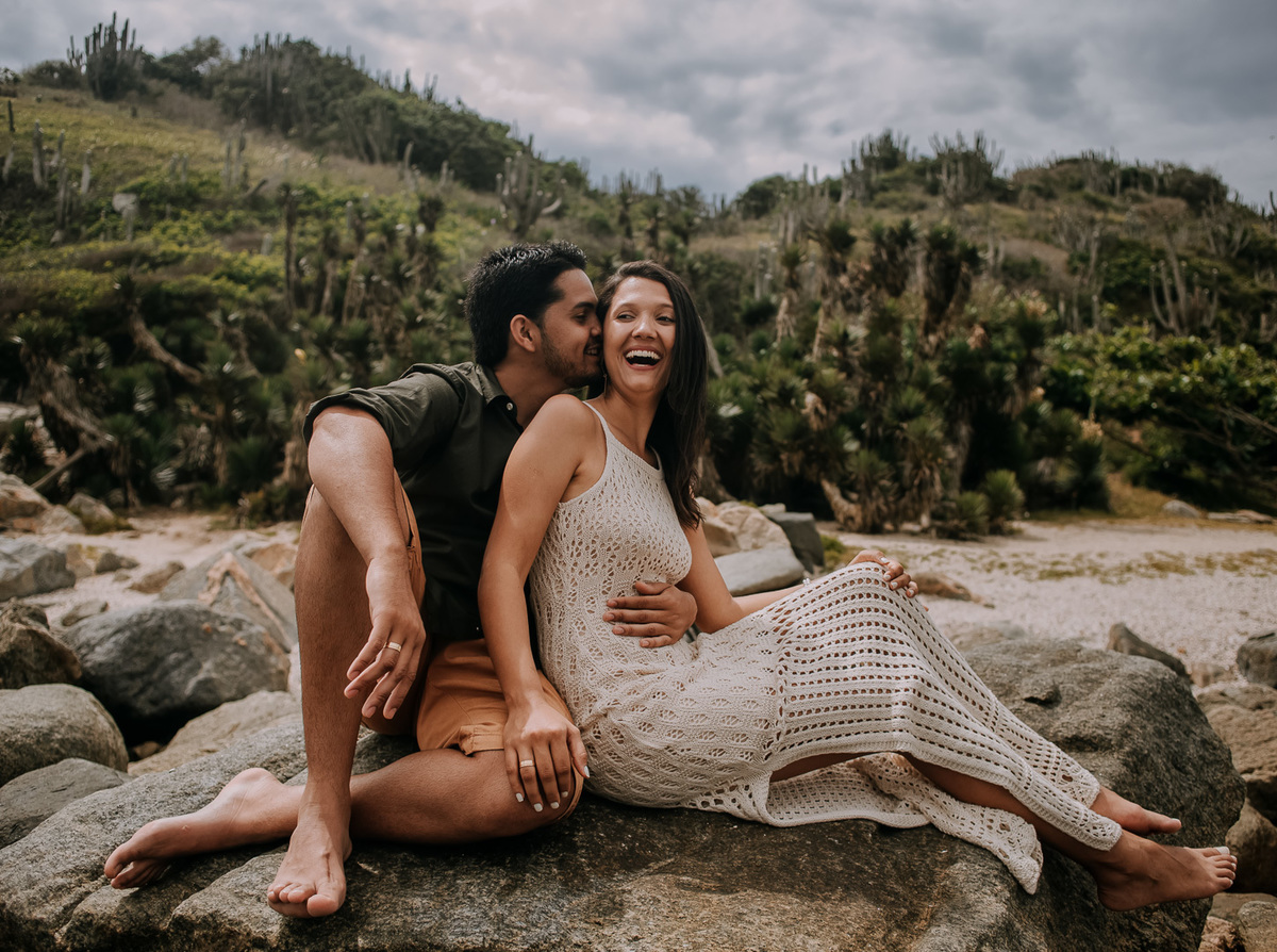 sentados na pedra da praia das conchas cabo frio noivos sorriem abraçados durante o ensaio de casal