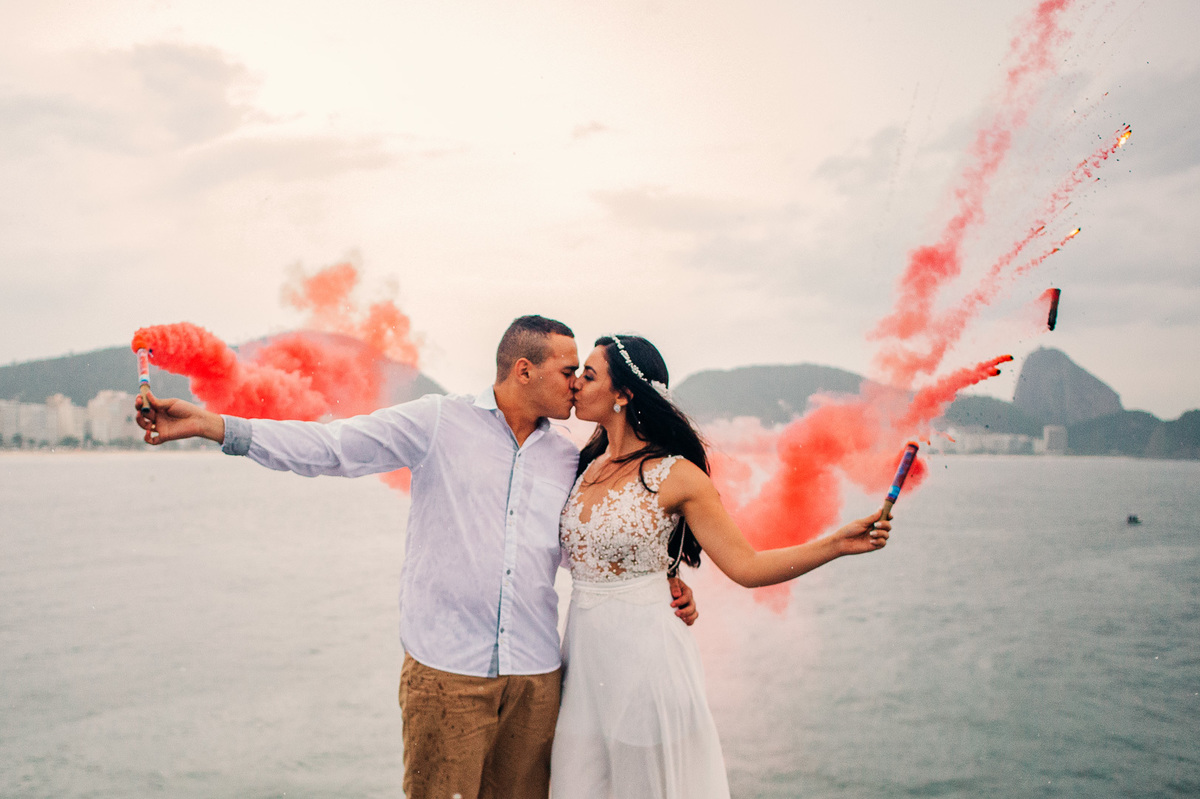 segurando sinalizadores de fumaça colorida casal se beijam no forte de copacabana com a praia de copacabana ao fundo
