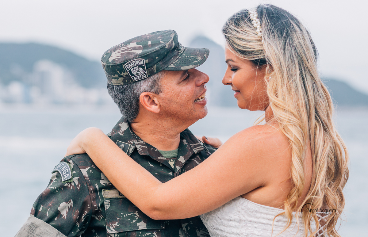fotografo de casamento rj, com os braços sobre o ombro do noivo, a noiva olha para o noivo de uma forma apaixonada na mureta do forte de copacabana