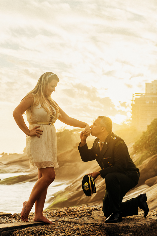 fotos ensaio pre casamento rj, noivo ajoelhado e beijando a mão da noiva durante o por do sol, nas ruínas do forte de copacabana por andre nogueira fotografia