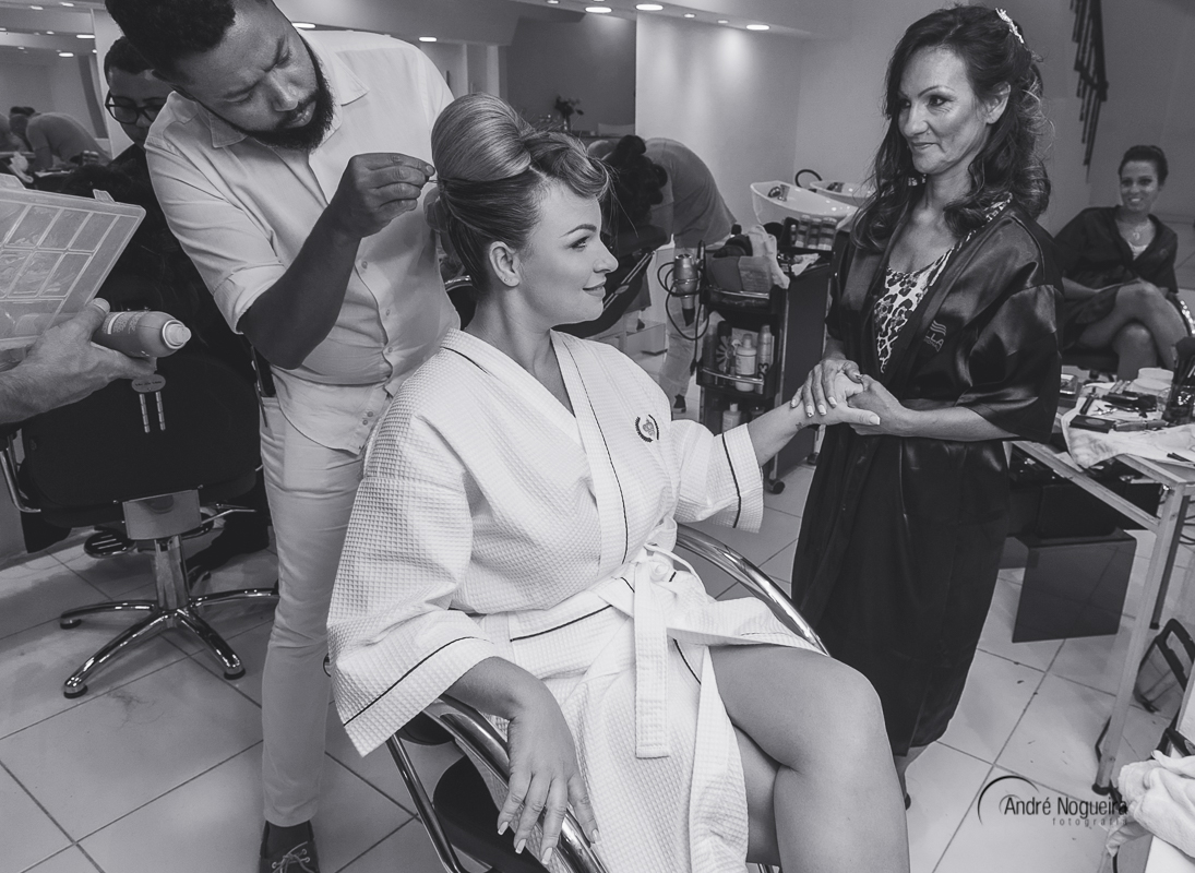 cabelereiro arrumando o cabelo da noiva, enquanto ela está de mãos dadas com a mãe e olhando uma para outra
