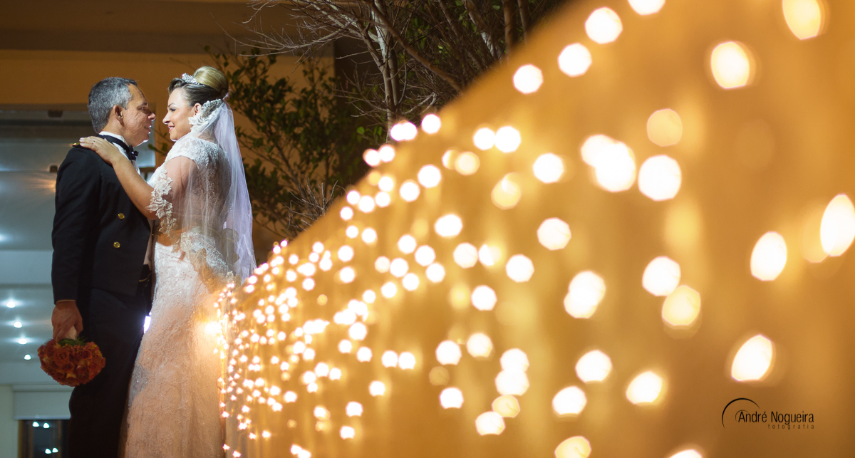 casal de frente um para outro se olhando apaixonadamente e o noivo segurando o boque de flores da noiva e encostados na mureta iluminada por lampadas amarelas