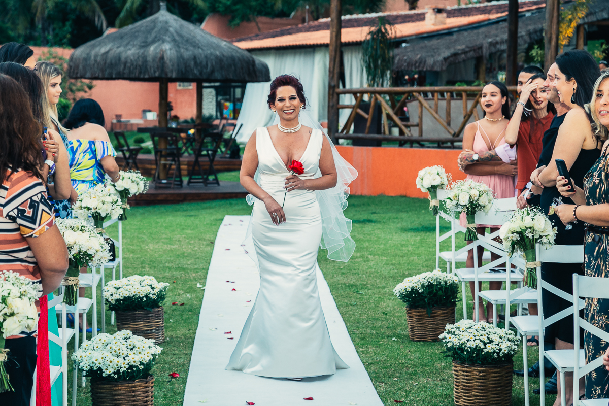 lá vem a noiva toda linda e sorridente, segurando uma rosa vermelha na mão e sendo fotografada por andré nogueira fotografia rumo ao altar para o seu casamento rj