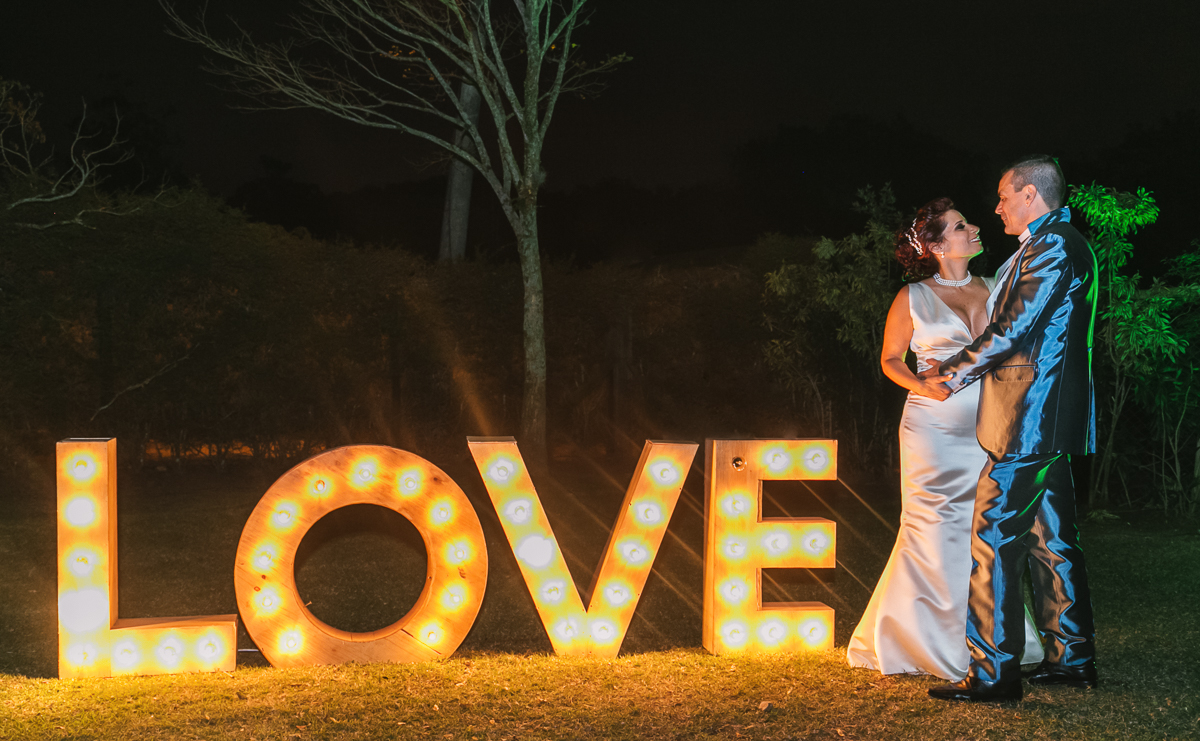 casamento no campo, noivos possando e se beijando no jardim da chácara do sapê ao lado de um letreiro iluminado com a palavra love, fotógrafo por andré nogueira fotografia