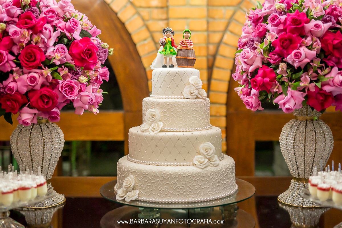 Bolo de casamento branco do casamento de Lívia e Thiago no Chalé Pampulha. Topo do bolo em biscuit bonecos chiquinha e chaves. Decoração com flores naturais rosas