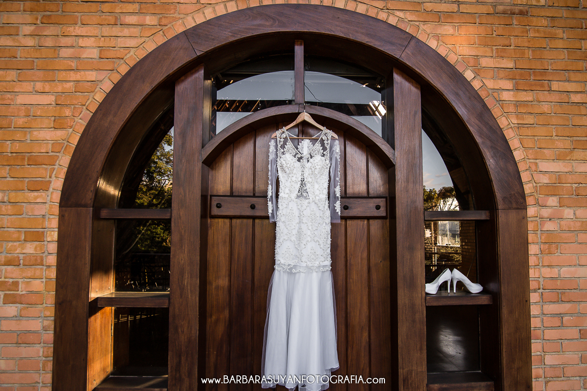 Making of da noiva, foto do vestido da noiva depenturado ao lado do sapato branco. Vestido da noiva dependurado na porta de madeira rústica do Chalé Pampulha.