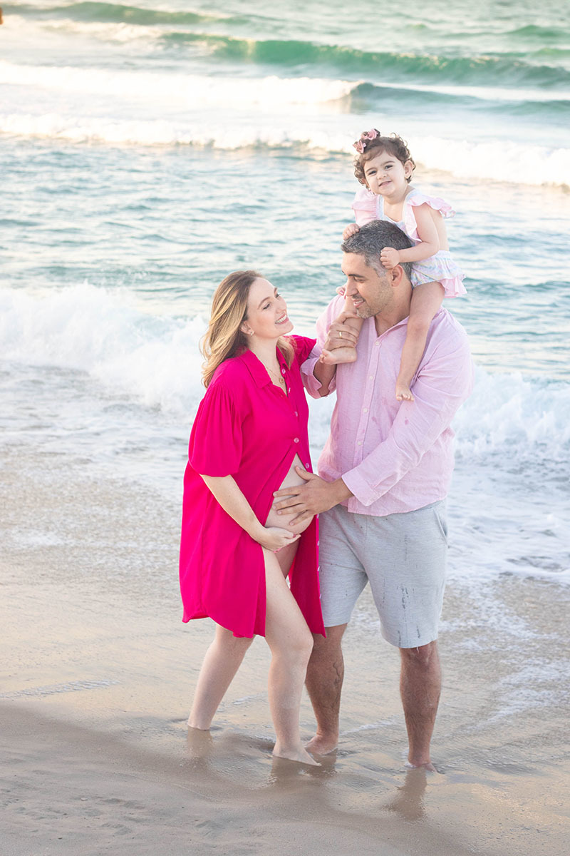 Casal sorrindo com a filha em ensaio gestante na Praia da Barra, Rio de Janeiro