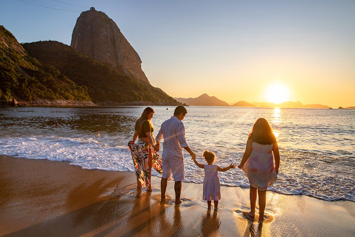 gestante, marido e duas filhas de costas, na beira do mar da Praia Vermelha de mãos dadas na areia da Praia Vermelha ao nascer do sol - ensaio gestante - nascer do sol - Rio de Janeiro - RJ.